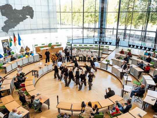 A group performs a dance in the centre of a round, modern auditorium with large windows. People sit around and watch, and flags are placed on the left. Outside there are trees and sunlight.
