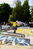 A young skateboarder in a yellow jacket and white helmet performs a trick on a rail in an outdoor skate park, with colourful graffiti and a crowd of spectators in the background.