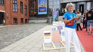 Eine Frau in blauem Hemd und weißer Hose steht vor einem Gebäude mit der Aufschrift "Industriemuseum", neben einem Display mit verschiedenen europäischen Flaggen und Ländernamen. Im Hintergrund sind mehrere Personen und ein roter Teppich zu sehen.