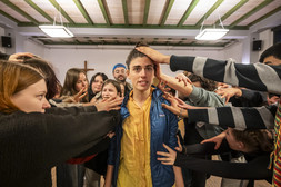 A person stands in the centre of a room and looks forward with a serious expression, while many people around them stretch out their hands to touch their head and shoulders. A wooden cross can be seen in the background.