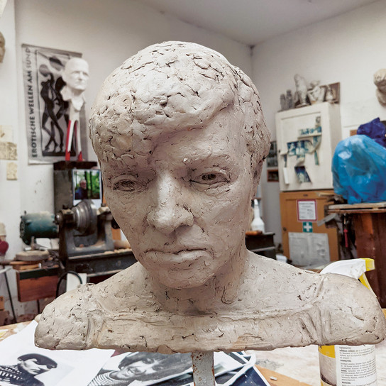 A clay bust of a person with a serious expression is displayed in an artist's studio. The background features other sculptures, sketches, and various art supplies.