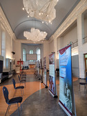 Geräumige, hohe Ausstellungshalle mit in der Mitte aufgereihten Schautafeln, umgeben von verstreuten Stühlen. Große Stoffleuchten hängen von der Decke, und Bogenfenster lassen Tageslicht herein.