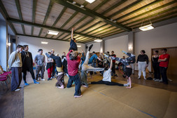 A group of people in casual clothes practise acro-yoga or group acrobatics in a spacious, well-lit room with a wooden floor and ceiling, with some forming a human pyramid and others watching or waiting their turn.
