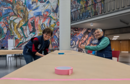 Zwei Menschen lächeln, während sie lange, mit braunem Papier bedeckte Tische mit Rollen von farbigem Klebeband vorbereiten. Ein großes, farbenfrohes Wandgemälde schmückt die Wand hinter ihnen in einem geräumigen Innenbereich.Foto: Josephine Hage 