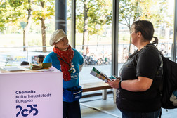 Ein Freiwilliger mit blauem Hemd und rotem Schal spricht mit einem Besucher, der eine Broschüre in der Hand hält, an einem Informationsstand der Kulturhauptstadt Europas 2025 in Chemnitz. Durch die Glasfenster sind Bäume und Sonnenlicht zu sehen.