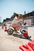 Ein kleines Kind mit einem gelben Helm fährt auf einem Balance-Bike an einem Verkehrskegel mit der Aufschrift "START" vorbei, in einem Außenbereich mit weiteren Kegeln und Gebäuden im Hintergrund.