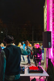 Ein DJ spielt Musik bei einer nächtlichen Veranstaltung im Freien, während die Menschen in der Nähe tanzen und sich unterhalten. Die Szene ist mit bunten Lichtern beleuchtet, darunter leuchtendes Pink an der Hauswand. Im Hintergrund sind Bäume und Freileitungen zu sehen.