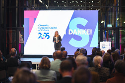 Eine Frau steht auf der Bühne und spricht zum Publikum. Hinter ihr ist eine große Leinwand zu sehen, auf der in fetten Lettern "Chemnitz Kulturhauptstadt Europas 2025" und "DANKE!" zu lesen ist. Der Raum ist hell erleuchtet und modern.
