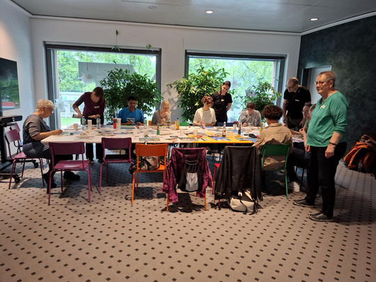 Eine Gruppe von Menschen sitzt und steht in einem hellen Raum mit großen Fenstern, Pflanzen und bunten Stühlen um einen mit Kunstbedarf bedeckten Tisch und ist mit einer kreativen Aktivität oder einem Workshop beschäftigt.