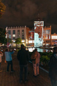 Menschen stehen nachts auf einem Weg und beobachten eine Lichtprojektion auf einen historischen Turm an einem Kanal, mit einem modernen Gebäude und Spiegelungen im Wasser im Hintergrund.