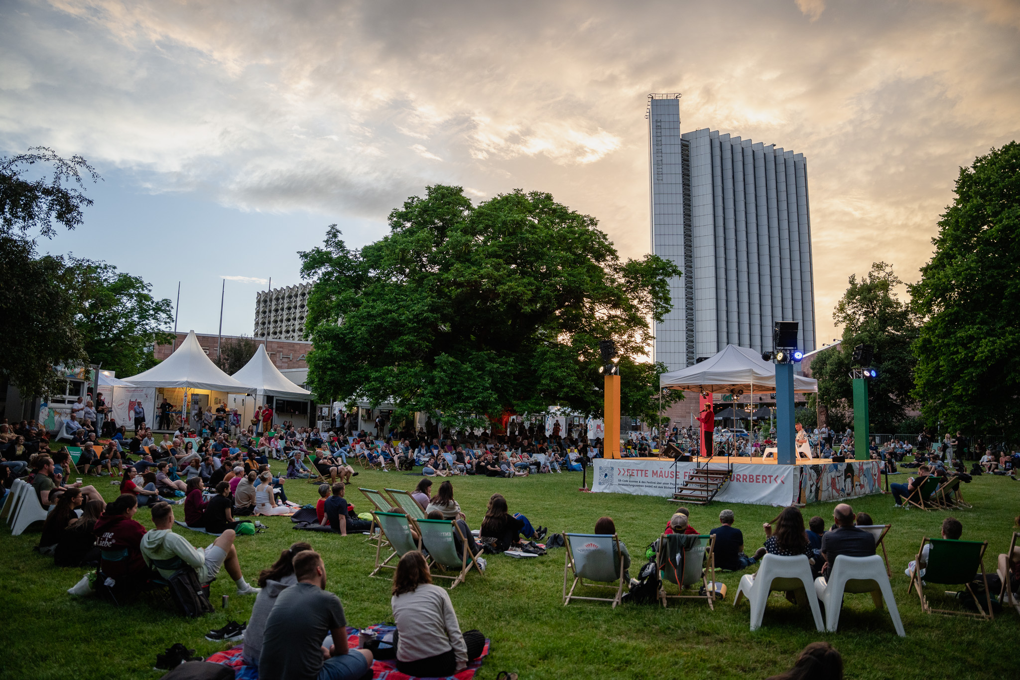 Eine große Menschenmenge sitzt im Gras und auf Stühlen bei einer Veranstaltung im Freien und beobachtet eine Bühnenvorführung bei Sonnenuntergang. Im Hintergrund sind Bäume, Zelte und ein hohes modernes Gebäude zu sehen.