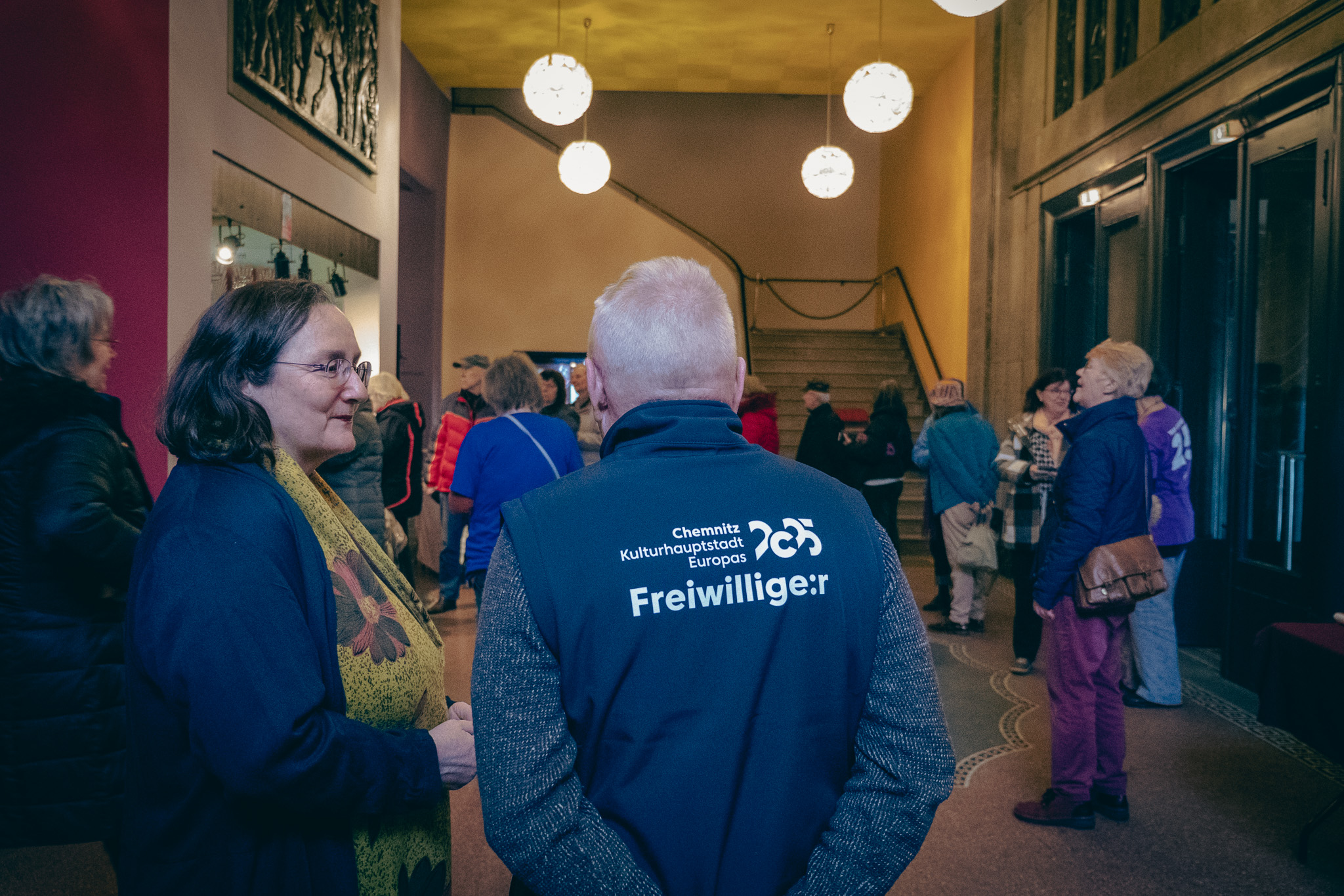 Eine Gruppe von Menschen steht in der Eingangshalle eines Gebäudes und unterhält sich. Eine Person trägt eine Weste mit der Aufschrift „Freiwillige:r“. Die Atmosphäre ist entspannt; andere sind versammelt oder gehen auf eine Treppe im Hintergrund zu. Kugelförmige Lichter hängen von der Decke.