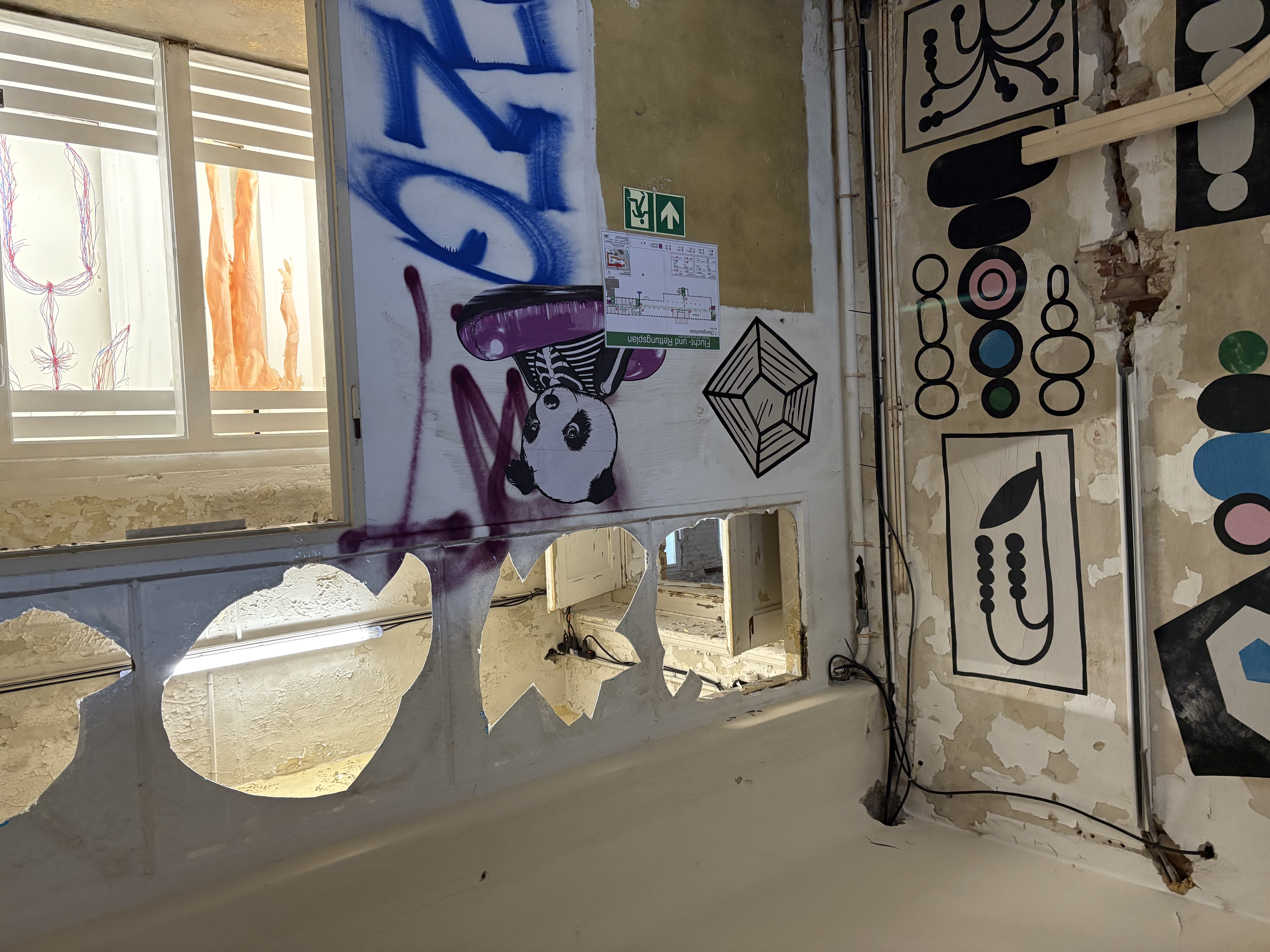 Ein Raum mit abblätternder Farbe, geometrischer Wandgestaltung und Graffiti mit einem Panda auf einem lila Autoscooter. Es gibt freiliegende Drähte, Notausgangsschilder und ein Fenster mit ausgeschnittenen Bögen über der Tür.