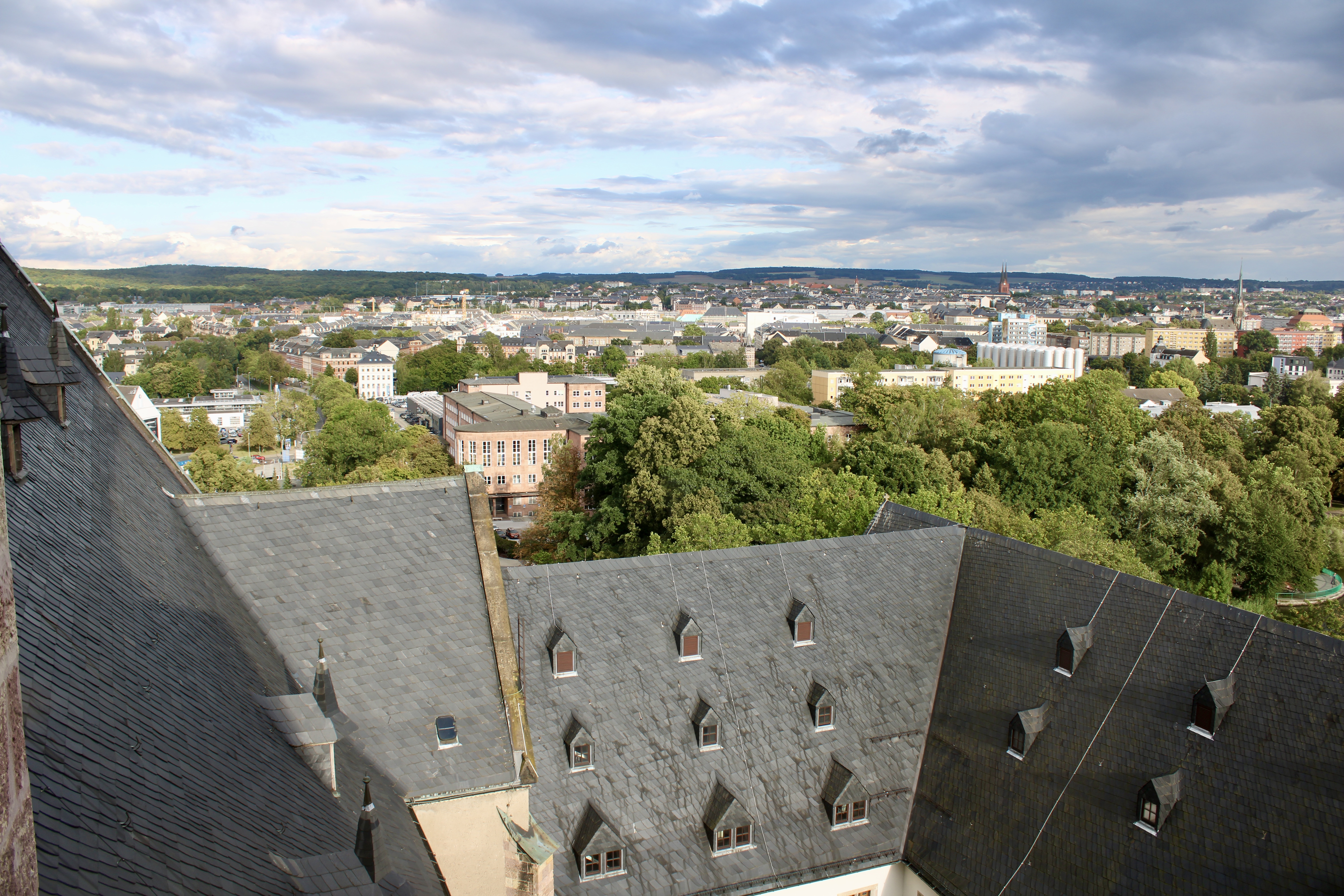 Eine Stadtansicht von oben zeigt schiefergedeckte Dächer mit dreieckigen Fenstern, grüne Bäume und eine weitläufige Stadt, die sich unter einem teilweise bewölkten Himmel in die Ferne erstreckt.