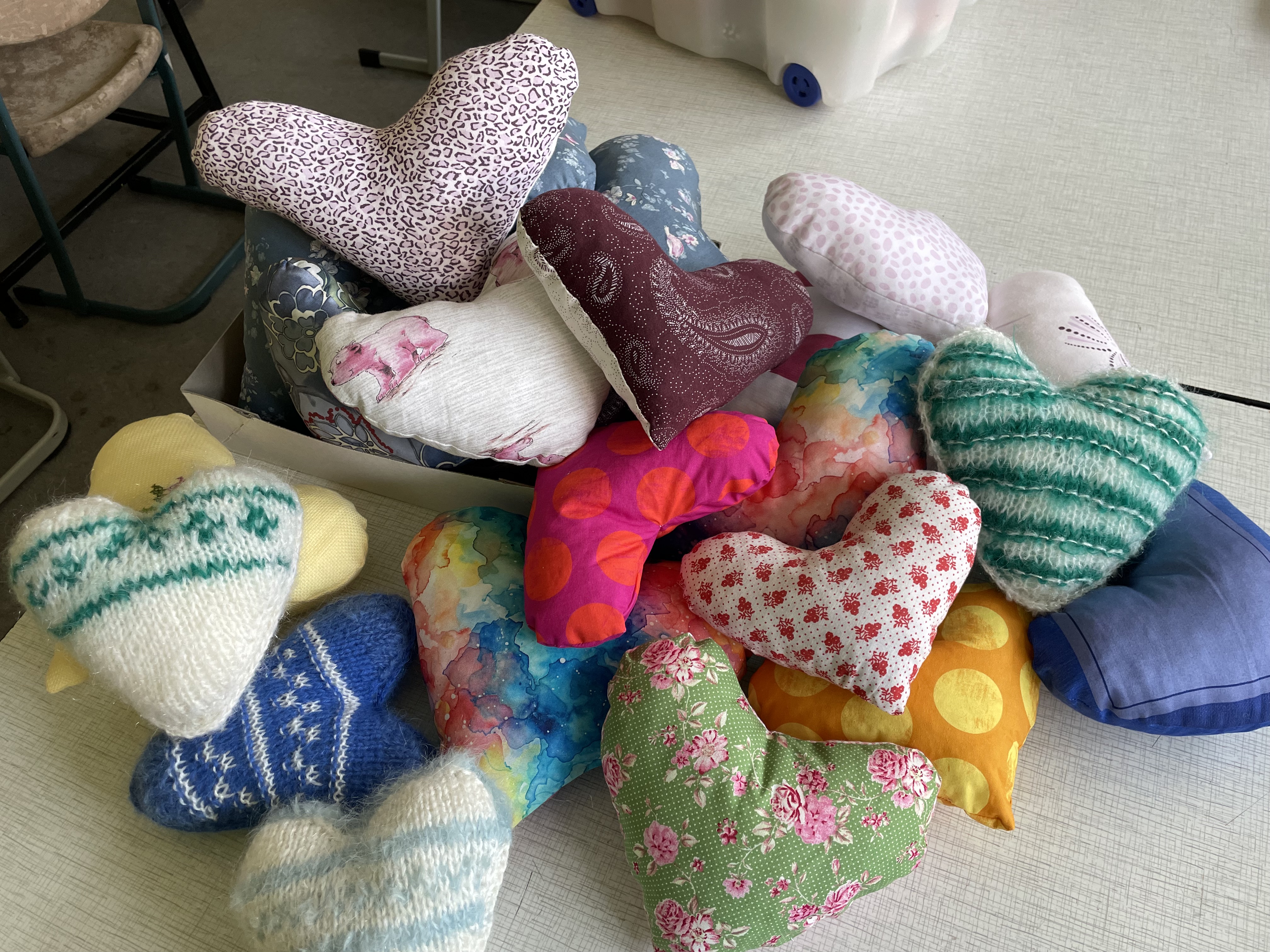 Eine Sammlung handgefertigter herzförmiger Kissen mit verschiedenen bunten Mustern und Stoffen, darunter Strick-, Blumen-, Tupfen- und Patchwork-Designs, angeordnet auf einem Tisch und in einer Schachtel.