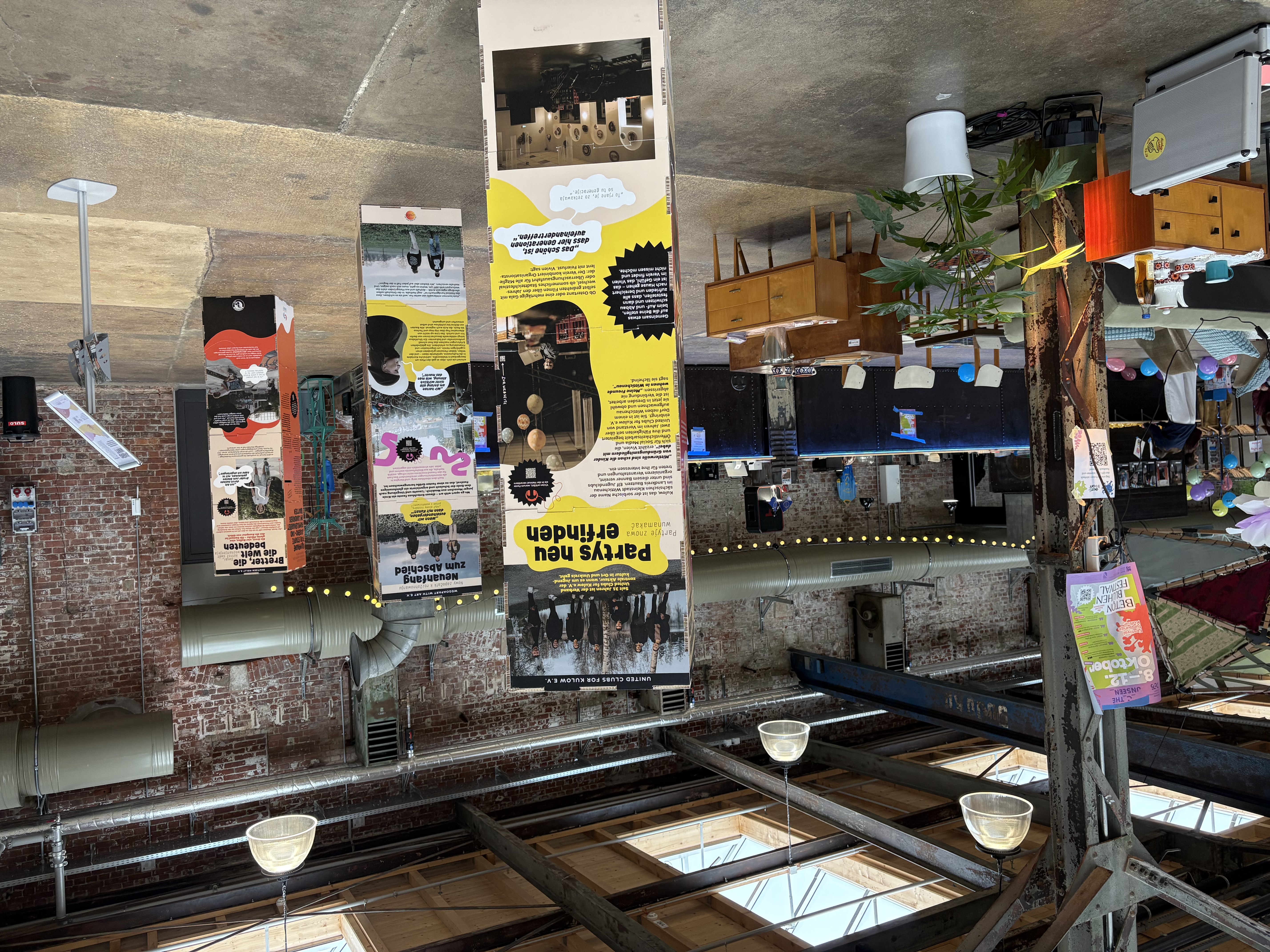 Ein überdachter Veranstaltungsraum mit freiliegenden Backsteinwänden und industriellen Balken bietet Informationstafeln, Retro-Möbel, Topfpflanzen und Menschen im Hintergrund. Das Sonnenlicht scheint durch die Oberlichter an der Decke.