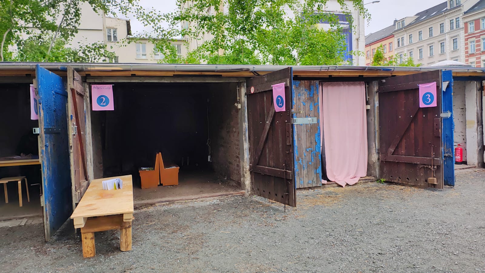 Drei Holzstände mit offenen Türen sind mit den Nummern 1, 2 und 3 gekennzeichnet. Stand 1 hat einen Tisch mit Papieren davor, Stand 2 ist leer und Stand 3 hat einen rosa Vorhang. Der Bereich ist mit Kiesboden und Gebäuden im Hintergrund bedeckt.