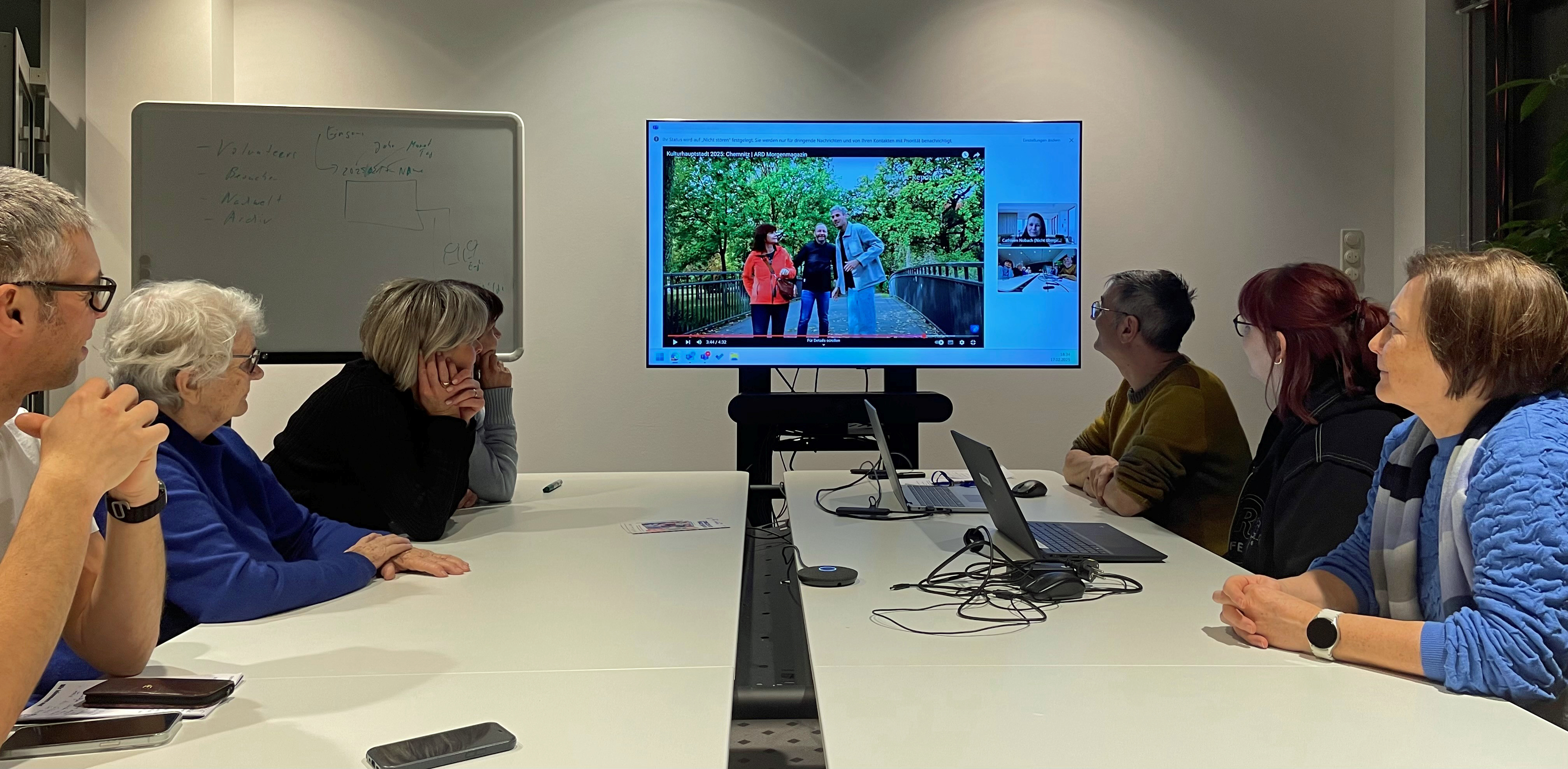 Sieben Personen sitzen an einem Konferenztisch und verfolgen eine Präsentation auf einem großen Bildschirm. Der Bildschirm zeigt ein Foto von einem Paar, das auf einer Brücke steht. Auf dem Tisch liegen ein Laptop und diverse Kabel. Im Hintergrund ist ein Whiteboard mit Notizen zu sehen.