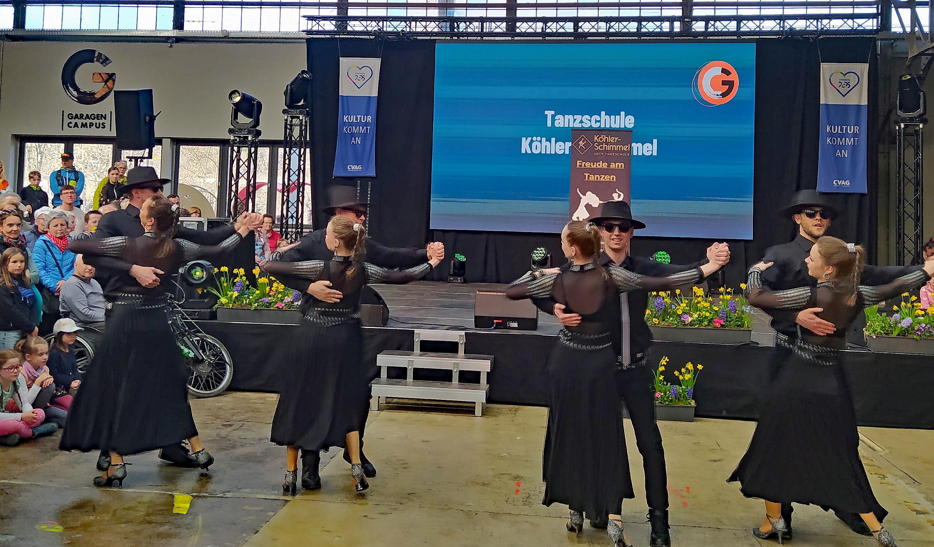 Vier Paare in schwarzen Outfits führen auf einer Indoor-Bühne einen Gesellschaftstanz auf. Zuschauer schauen zu, auf einer Leinwand dahinter ist „Tanzschule Köhler-Schimmel“ zu sehen. Der Saal ist mit Blumen und Bannern geschmückt.