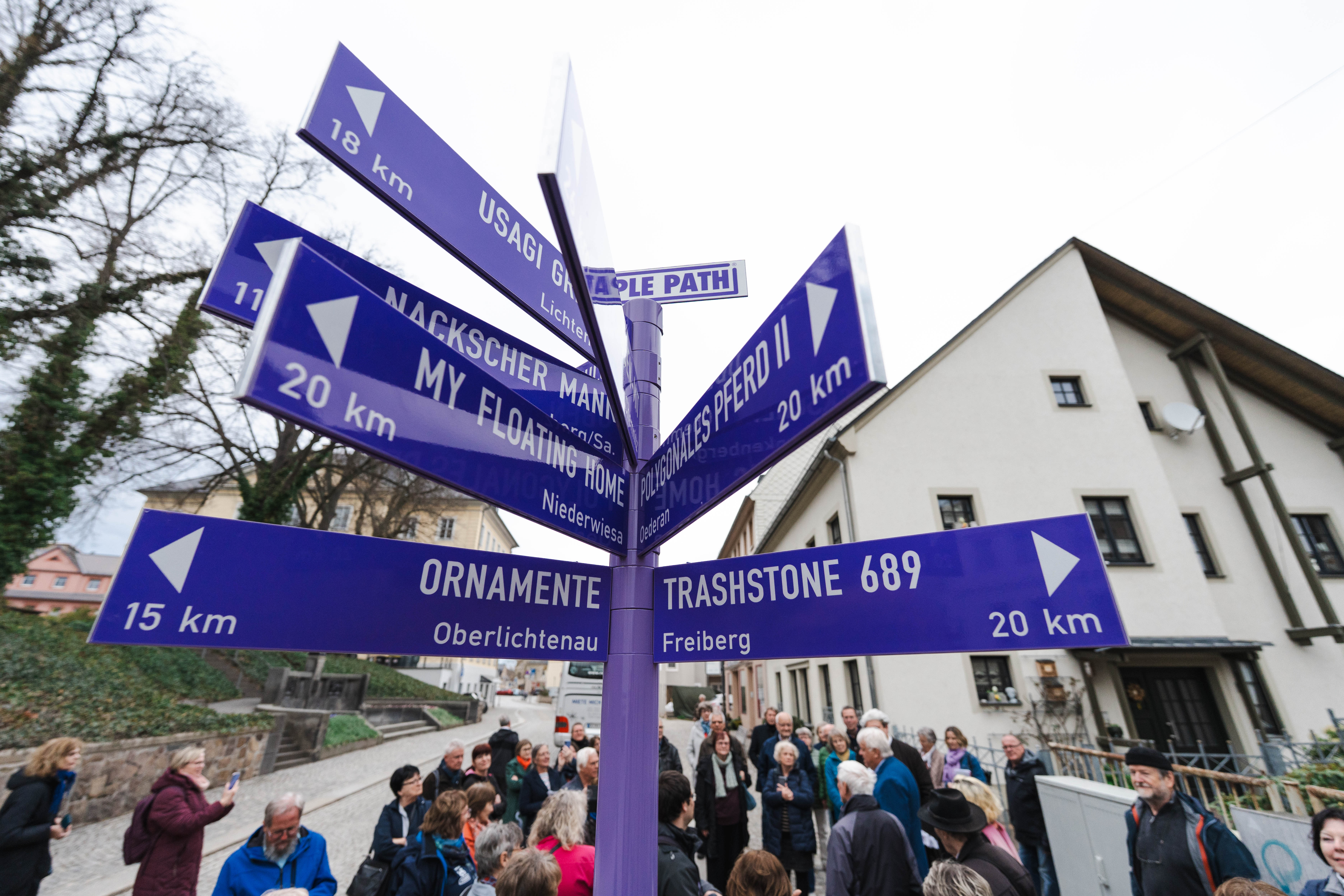 Eine Gruppe von Menschen steht um einen violetten Wegweiser am PURPLE PATH in Hainichen mit mehreren Pfeilen, die in verschiedene Richtungen weisen und jeweils mit Zielnamen und Entfernungen in Kilometern beschriftet sind.